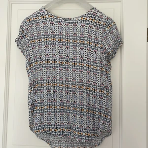 Mönstrad blus från H&M - Säljer en blus från H&M i storlek 38 med korta ärmar och rund hals. Blusen har ett färgglatt geometriskt mönster i blått, svart, vitt, rött och gult. Tillverkad i ett lätt och luftigt material, perfekt för varmare dagar.