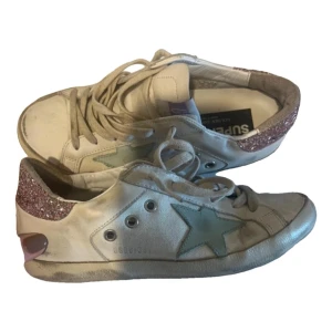 Vita sneakers från Golden Goose med glitterdetaljer - Golden Goose Super-Star sneakers i vitt skinn med ljusblå stjärna på sidan och rosa glitter på hälen. Skorna har snörning och platt sula, samt en klassisk rund tå. Perfekta för dig som gillar unika detaljer och streetstyle. Äkthetsbevis finns