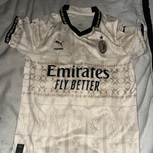 AC Milan bortatröja - Säljer en vit AC Milan bortatröja från Puma med svarta detaljer och guldfärgade mönster. Tröjan har korta ärmar, svart krage med mönster och klubbmärke på bröstet. Perfekt för dig som älskar fotboll och vill visa ditt stöd för Milan.