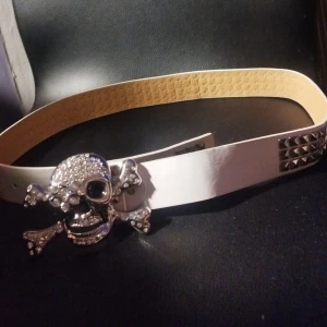 Vitt bälte med dödskalle-spänne och nitar - Säljer ett vitt bälte med coolt dödskalle-spänne i silver och glittriga detaljer. Bältet har fyrkantiga silverfärgade nitar längs hela bandet. Perfekt accessoar för att sticka ut och ge outfiten edge.