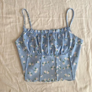Blommig blå croppad linnetopp från SHEIN - Supersöt ljusblå croppad linnetopp från SHEIN med små vita och rosa blommor. Toppen har smala axelband och lätt rynk vid bysten för en feminin look. Perfekt för varma dagar och enkel att matcha med jeans eller kjol.