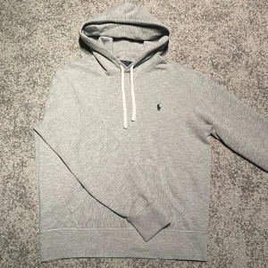 Ralph Lauren hoodie  - Snygg grå hoodie från Polo Ralph Lauren med klassisk broderad logga på bröstet. Tröjan har huva med vita snören och en stor magficka framtill. Perfekt för en avslappnad stil.