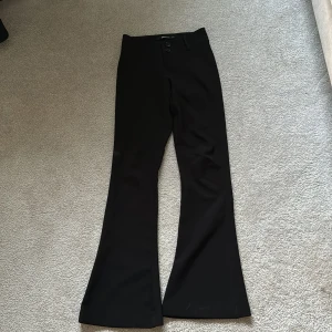 Svarta bootcut kostymbyxor från Gina Tricot - Säljer ett par svarta kostymbyxor från Gina Tricot i bootcut-modell. Byxorna har två knappar framtill, bälteshällor och fickor med lock bak. Perfekta för en stilren look.