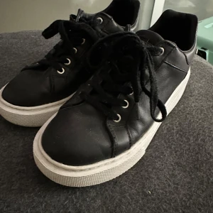 Svarta sneakers från Guess - Säljer ett par svarta sneakers från Guess med vita sulor och diskret logotypmönster på sidan. Skorna har snörning och metallfärgad G-detalj på hälen. Perfekta för en stilren vardagslook. Säljes inte längre. Kom gärna med bud!