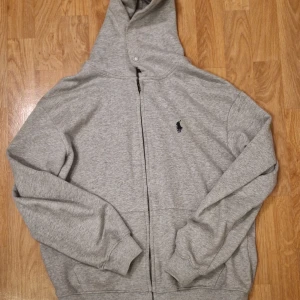 Grå hoodie från Polo Ralph Lauren - Säljer en klassisk grå hoodie från Polo Ralph Lauren med dragkedja och broderad logga på bröstet. Tröjan har långa ärmar, huva och ribbade muddar. Perfekt för en avslappnad stil.