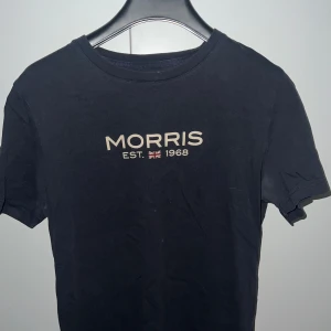 Svart t-shirt från Morris - Svart t-shirt från Morris med tryckt text 'MORRIS EST. 1968' och en liten brittisk flagga på bröstet. Klassisk rund hals och korta ärmar. Perfekt för en stilren och enkel look.