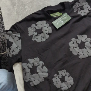 Svart Denim Tears Shirt - Passar som M/L. Säljer en svart t-shirt från Denim Tears med stora vita blommönster tryckta på bröstet och axlarna. Tröjan har rund halsringning och är i mjuk bomull. Perfekt för dig som vill sticka ut med en unik design.