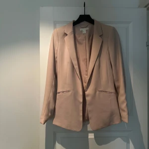 Ljusrosa/Beige kavaj från H&M - Elegant ljusrosa/beige kavaj från H&M med klassisk krage och två fickor framtill. Långärmad modell med rak passform som ger en sofistikerad och feminin look. Tillverkad i ett mjukt, slätt material som ger ett stilrent intryck. Storlek 34