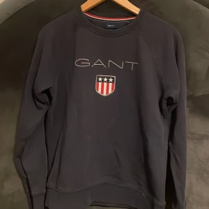 Svart sweatshirt från GANT med broderad logga - Säljer en svart sweatshirt från GANT med broderad logga och sköld med stjärnor och ränder på bröstet. Tröjan har rund halsringning och långa ärmar. Klassisk och stilren design med mjukt material.