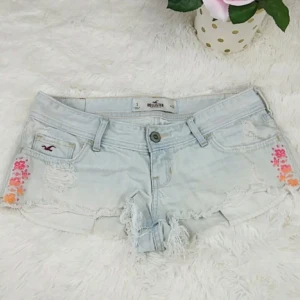 Lågmidjade jeansshorts - Ljusblå jeansshorts från Hollister med slitna detaljer och broderade blommor i rosa, rött och orange på sidorna🩷skriv innan ni köper!