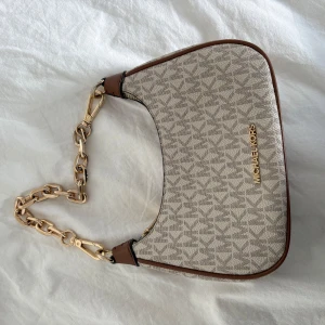 Beige axelväska från Michael Kors med kedjedetalj - Säljer en snygg beige axelväska från Michael Kors med guldiga kedjedetaljer och dragkedja. Väskan säljs inte längre då modellen är slutsåld. Nypris 3500, pris kan diskuteras vid snabb affär. 