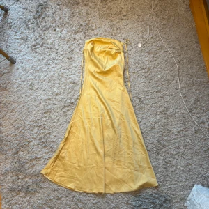 Gul långklänning med smala axelband - Säljer en snygg gul långklänning med smala axelband och lätt glansigt tyg. Klänningen har en elegant, figurnära passform upptill och faller ut nertill. Perfekt för sommarens tillfällen!
