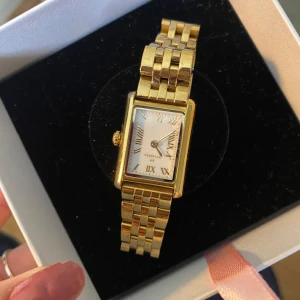 Mockberg klocka - Säljer en elegant guldfärgad klocka från Mockberg med fyrkantig vit urtavla och romerska siffror. Klockan har ett klassiskt länkarmband i metall och levereras med extra länkar för justering av storlek.