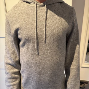 Grå stickad hoodie - Säljer en enkel och stilren grå stickad hoodie med huva och dragsnören. Perfekt för dig som gillar en avslappnad look. Tröjan har långa ärmar och är mjuk och bekväm att bära.