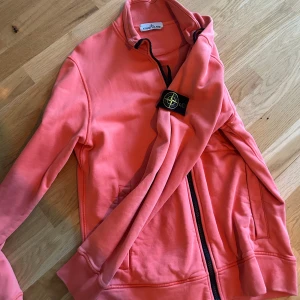 Korallfärgad zip-up från Stone Island - Säljer en snygg korallfärgad zip-up från Stone Island med svart dragkedja och klassisk logotyp på ärmen. Jackan har hög krage och två fickor framtill. Perfekt för dig som vill ha en stilren och bekväm tröja med streetkänsla.