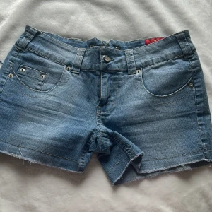 Blå jeansshorts från Hongma - Säljer ett par klassiska blå jeansshorts från Hongma med fem fickor, bälteshällor och råa kanter nertill. Shortsen har knapp- och dragkedjestängning samt dekorativa nitar vid fickorna. Perfekta för varma dagar!