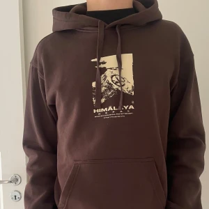 Brun hoodie med shysst tryck - Säljer en brun hoodie med tryck av Himalaya och text på bröstet. Tröjan har huva med dragsko och en stor magficka framtill. Perfekt för en avslappnad stil.