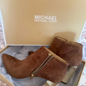 Bruna mocka boots från Michael Kors - Säljer ett par snygga bruna boots i mocka från Michael Kors. Skorna har guldfärgade dragkedjor på sidan och en klack som ger extra höjd. Perfekta för dig som vill ha en stilren och trendig look. Använda endast 1 gång 