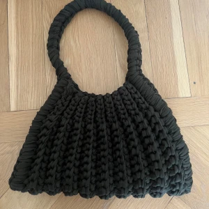 Svart stickad axelväska från OJWA - Säljer en svart stickad axelväska från OMA. Väskan har ett unikt, grovstickat mönster och ett mjukt, vadderat handtag som gör den bekväm att bära. Perfekt för dig som vill ha en trendig accessoar med personlig touch.