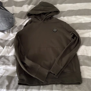 Mörkgrön hoodie från J.Lindeberg - Snygg mörkgrön hoodie från J.Lindeberg med klassisk huva och en diskret logotyp på bröstet. Tröjan har långa ärmar och en stor magficka framtill. Perfekt för en avslappnad och stilren look.