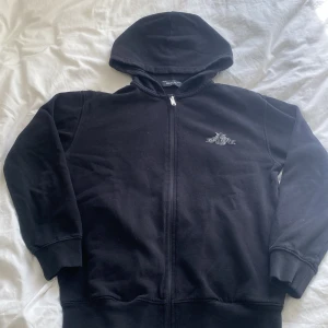 Svart hoodie med dragkedja från Replay - Svart hoodie från Replay med dragkedja framtill och stor logga i vitt på ryggen. Tröjan har huva och ribbade muddar vid ärmslut och nederkant. Perfekt för en avslappnad och cool stil.