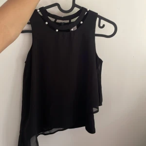 Svart blus  med pärldetaljer - Säljer en svart blus med pärldetaljer runt halsringningen. Toppen är i ett tunt, flowy material och har en dragkedja och en knapp bak vid nacken. Säljer den då den är för liten, storlek 134/140