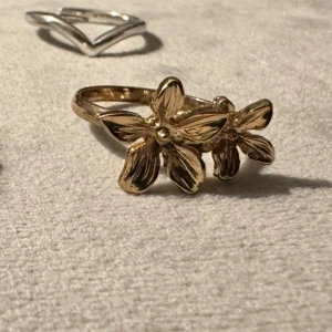 Guldfärgad ring med blommor - Säljer en snygg guldfärgad ring med två detaljerade blommor framtill. Ringen har en elegant och unik design som sticker ut och passar perfekt för dig som gillar smycken med naturinspirerade detaljer. Tror storleken är typ 54, vet ej exakt