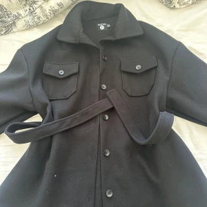 Svart kappa med bälte från Boohoo - Säljer en svart kappa från Boohoo med bälte i midjan och stora bröstfickor med knappar. Jackan har klassisk krage och knäppning framtill. Perfekt för dig som vill ha en stilren och enkel look. Lite stor i storleken så den passar även på M och till och med L. Aldrig använd.