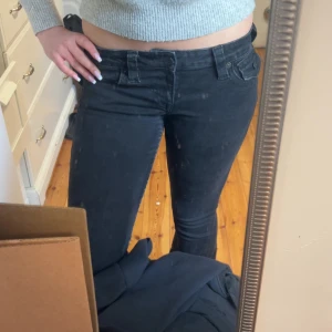 Svarta True Religion Joey Low Rise Flare jeans - Säljer ett par svarta True Religion Joey Low Rise Flare jeans med snygga fickdetaljer och knappar bak. Byxorna har låg midja och utställda ben, klassisk True Religion-logga på bakfickan och grova sömmar. Perfekta för dig som gillar en cool och tidlös jeansstil.