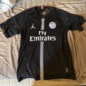 Psg tröja - Säljer en svart PSG fotbollströja med vita detaljer och Fly Emirates-tryck. Tröjan har korta ärmar, PSG-logga på bröstet och en randig detalj i mitten. Tillverkad i lätt och ventilerande Dri-FIT-material.