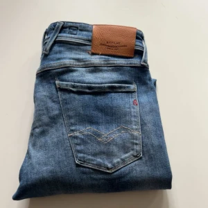 Replay Anbass slim fit jeans - Snygga blå Replay Anbass jeans i slim fit med slitna detaljer och klassisk femficksdesign. Jeansen har en läderpatch bak i midjan och tydliga slitningar framtill för en trendig look. Perfekta för dig som gillar en smalare passform och stilren denim.