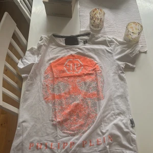 Vit t-shirt med dödskalle från Philipp Plein - Säljer en vit t-shirt från Philipp Plein med en stor glittrig dödskalle i orange och silver på framsidan. T-shirten har rund halsringning och korta ärmar. Märkeslogga syns både i nacken och på insidan av kragen.