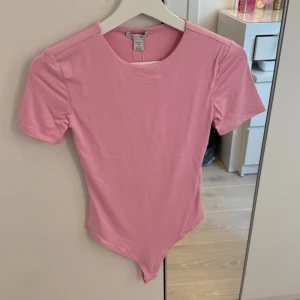 Rosa body från New Yorker - Säljer en rosa body från New Yorker med rund halsringning och korta ärmar. Perfekt att styla till jeans eller kjol för en enkel och snygg look. Materialet är mjukt och stretchigt för en bekväm passform.