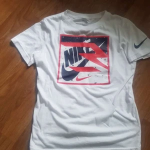 Vit t-shirt med grafiskt Nike-tryck - Vit t-shirt från Nike med stort grafiskt tryck i rött, blått och svart på bröstet samt Nike-logga på ärmen. Klassisk passform och rund hals. Tillverkad i en mjuk bomulls- och polyestermix.