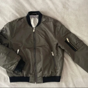 Olivgrön bomberjacka från Zara Basic - Snygg olivgrön bomberjacka från Zara Basic med svarta muddar och dragkedjor i silver. Jackan har två fickor framtill och en ärmficka med dragkedja. Perfekt för dig som vill ha en stilren och trendig look.