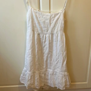 Vit sommarklänning med volang och öppen rygg - Söt vit klänning med smala axelband, volang nertill och öppen rygg med knyt. Perfekt för varma dagar och midsommar. Tillverkad i ett lätt och luftigt material som känns skönt mot huden.