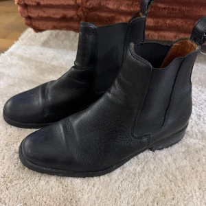 Svarta chelsea boots i skinn - Klassiska svarta chelsea boots i skinn med elastiska paneler på sidorna. Snygg och tidlös modell som passar till många olika outfits. Rund tå och låg platt sula för en bekväm känsla hela dagen. Storlek 37 men stor i storleken så den är som en 38. 