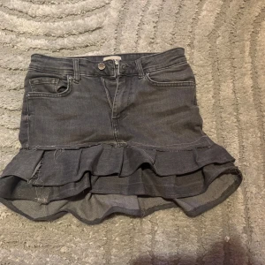 Grå jeanskjol med volang - Säljer en grå jeanskjol med volangdetalj nedtill. Kjolen  har knapp och dragkedja framtill samt råa kanter vid volangen för en cool look. Perfekt för dig som gillar en trendig och avslappnad stil.