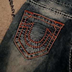 Blå jeans True Religion - Säljer ett par blå jeansbyxor från True Religion med tydliga vita och orange kontrastsömmar och ikoniska bakfickor. Modellen heter Jackson och har raka ben. Klassisk femficksdesign och lågmidjad passform.
