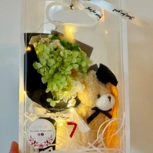 Presentförpackning med nalle och blombukett - Söt presentförpackning med en mjuk nallebjörn som bär studentmössa och svart rosett, samt en konstgjord blombukett i grönt och vitt. Perfekt som gåva till studenten.