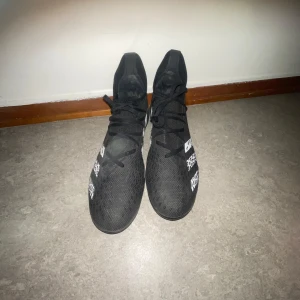 Svarta Adidas fotbollsskor med vita detaljer predators - Säljer ett par svarta Adidas fotbollsskor med vita ränder och textdetaljer. Skorna har snörning och en högre strumpliknande krage för extra stöd. Perfekta för dig som vill ha stilrena och funktionella skor på planen.