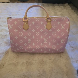 Rosa monogram handväska speedy 40 - Säljer en rosa handväska med klassiskt monogrammönster i vitt. Väskan är en stor weekendbag och har guldfärgade metalldetaljer, handtag i beige läder och dragkedja upptill. Perfekt för dig som vill ha en lyxig accessoar med ikonisk design.