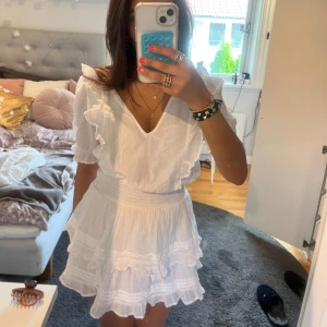 Vit volangklänning med puffärmar - Supersöt vit klänning med volanger och puffiga kortärmar. Klänningen har v-ringning, markerad midja och flera lager volang i kjolen. Perfekt för sommarens alla tillfällen och ger en romantisk känsla.