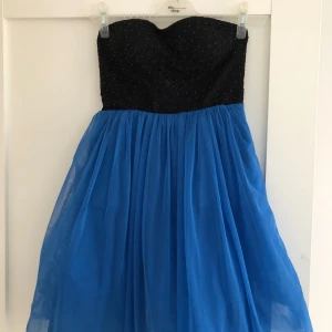 Blå och svart axelbandslös klänning - Säljer en snygg axelbandslös klänning med svart glittrig överdel och vid blå kjol i tyll. Perfekt för dig som vill sticka ut med en färgstark look. Kjolen är luftig och ger en fin siluett.