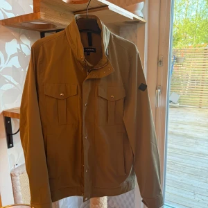 Olivgrön jacka från J Lindberg (Bailey) - Snygg olivgrön jacka från Lindbergh med hög krage och knappar framtill. Jackan har två bröstfickor med lock och knapp samt diskret logotyp på ärmen. Perfekt för vår och höst.