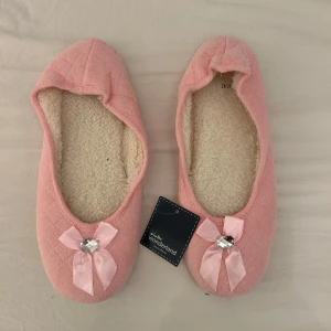 Rosa tofflor med rosett från Winter Wonderland - Mysiga rosa tofflor från Winter Wonderland med mjukt vitt foder. Dekorerade med en söt rosett och en glittrig sten framtill. Perfekta att ha inomhus när det är kallt. I strl 41/42