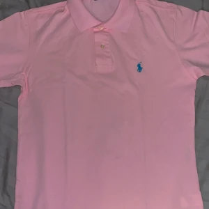 Polo Ralph Lauren piké - Säljer en ljusrosa pikétröja från Polo Ralph Lauren med klassisk krage och två knappar. Custom slim fit-modell med blå broderad logga på bröstet. Perfekt för en stilren och avslappnad look.