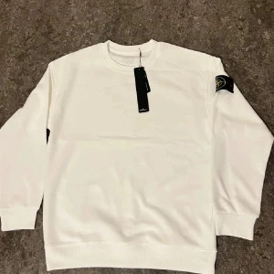 Vit sweatshirt från Stone Island - Säljer en stilren vit sweatshirt från Stone Island med klassisk logotyp-patch på vänster ärm. Tröjan har rund halsringning och ribbade muddar vid ärmslut och nederkant. Perfekt för en clean och avslappnad look.