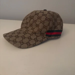 Beige keps från Gucci  - Säljer en beige keps från Gucci med klassiskt GG-monogram över hela kepsen. Kepsen har en böjd skärm, justerbar rem baktill i brunt och en röd-blå randig detalj på sidan. Perfekt för dig som gillar lyxiga accessoarer.
