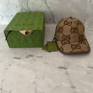 Beige GG keps från Gucci - Säljer en beige keps från Gucci med klassiskt GG-mönster i brunt. Kepsen har böjd skärm och justerbar rem baktill. Kommer med originalkartong och dustbag. Perfekt för dig som gillar lyxiga accessoarer.